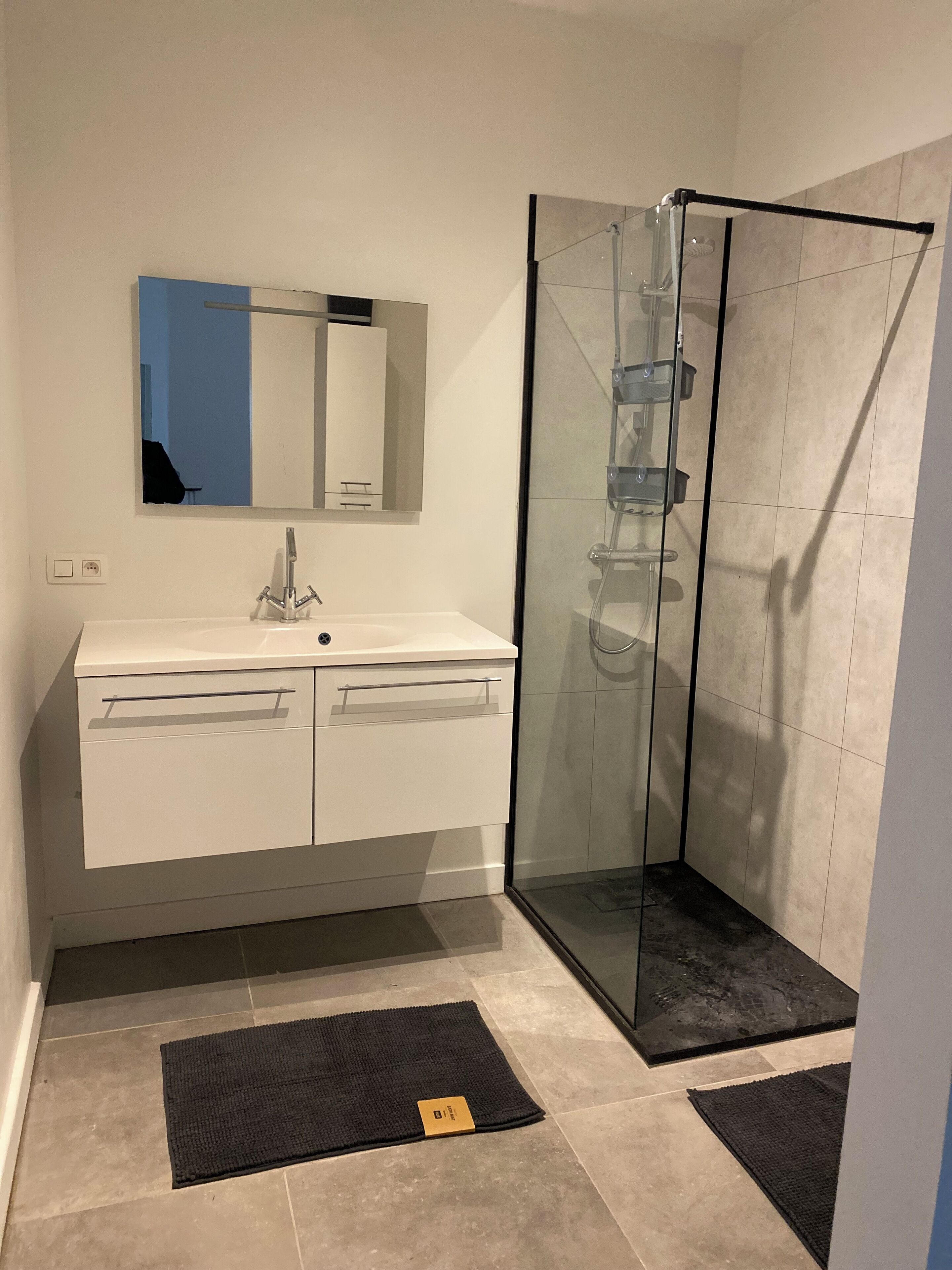 A modern bathroom with a glass-enclosed shower, a white vanity with a mirror, and two black mats on the floor.
