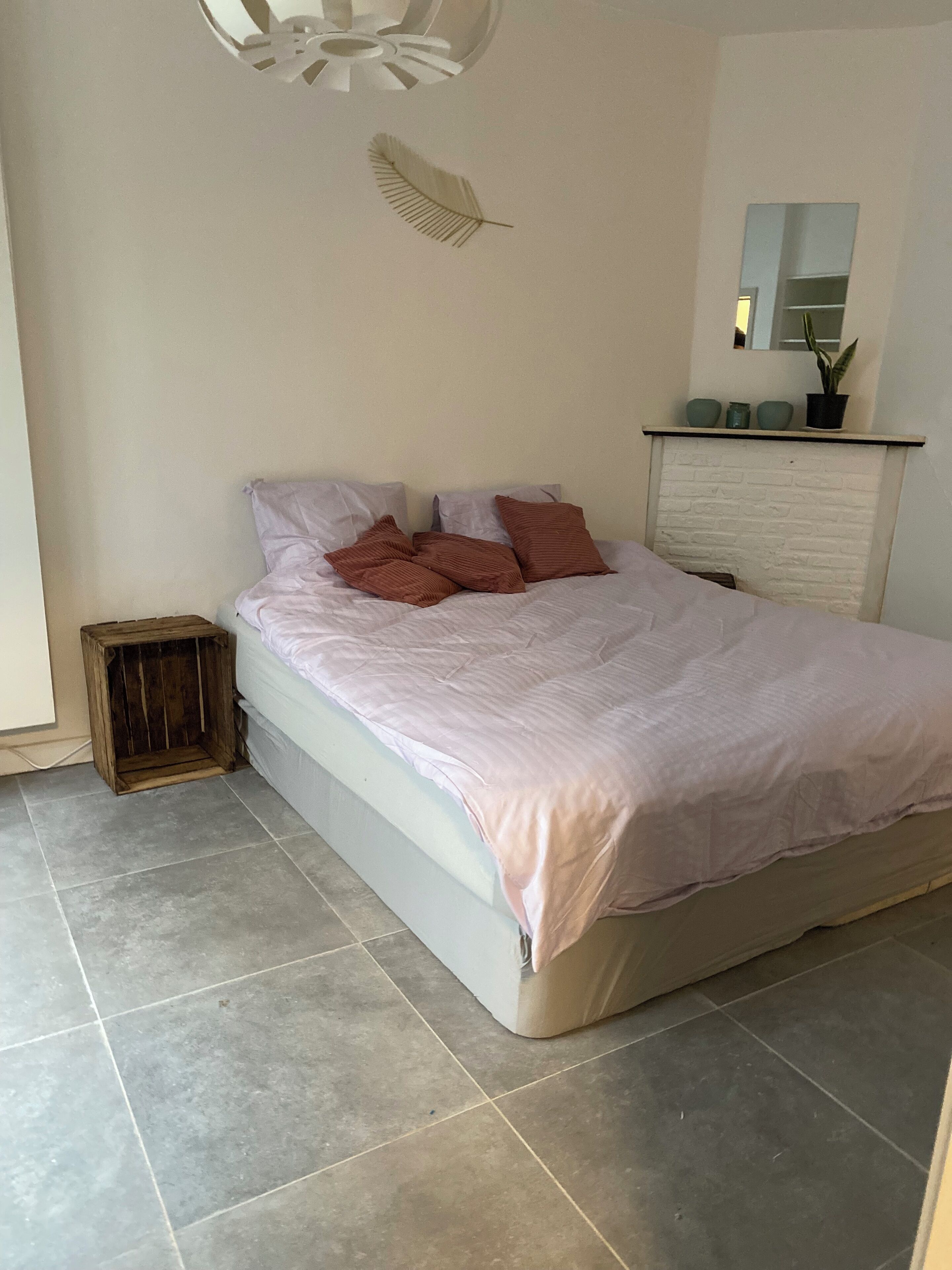 Un dormitorio con cama, almohadas, un taburete de madera, una chimenea de ladrillo blanco y un espejo en la pared.