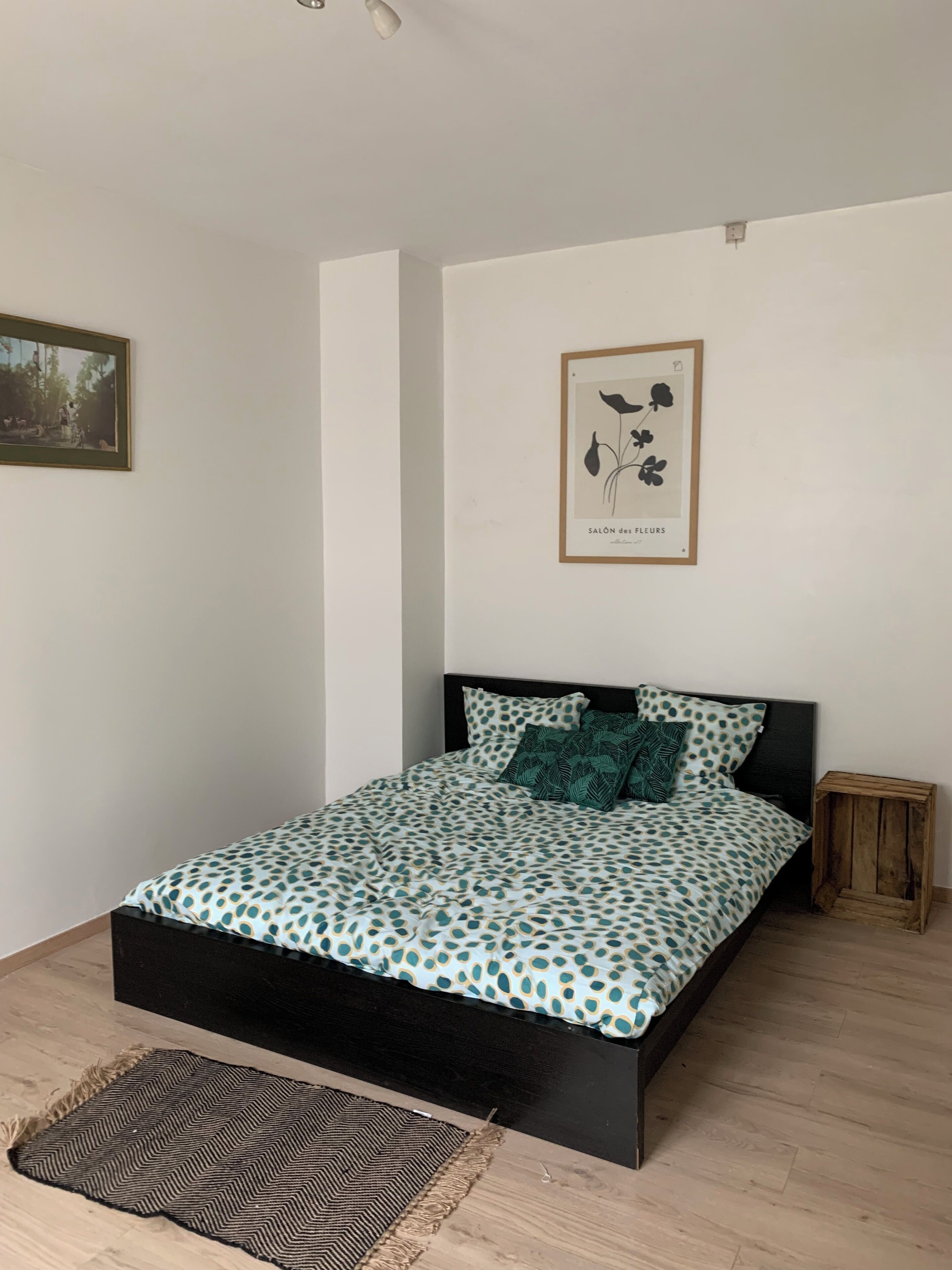 A bedroom with a bed, a small wooden side table, a framed botanical print, and a patterned rug.