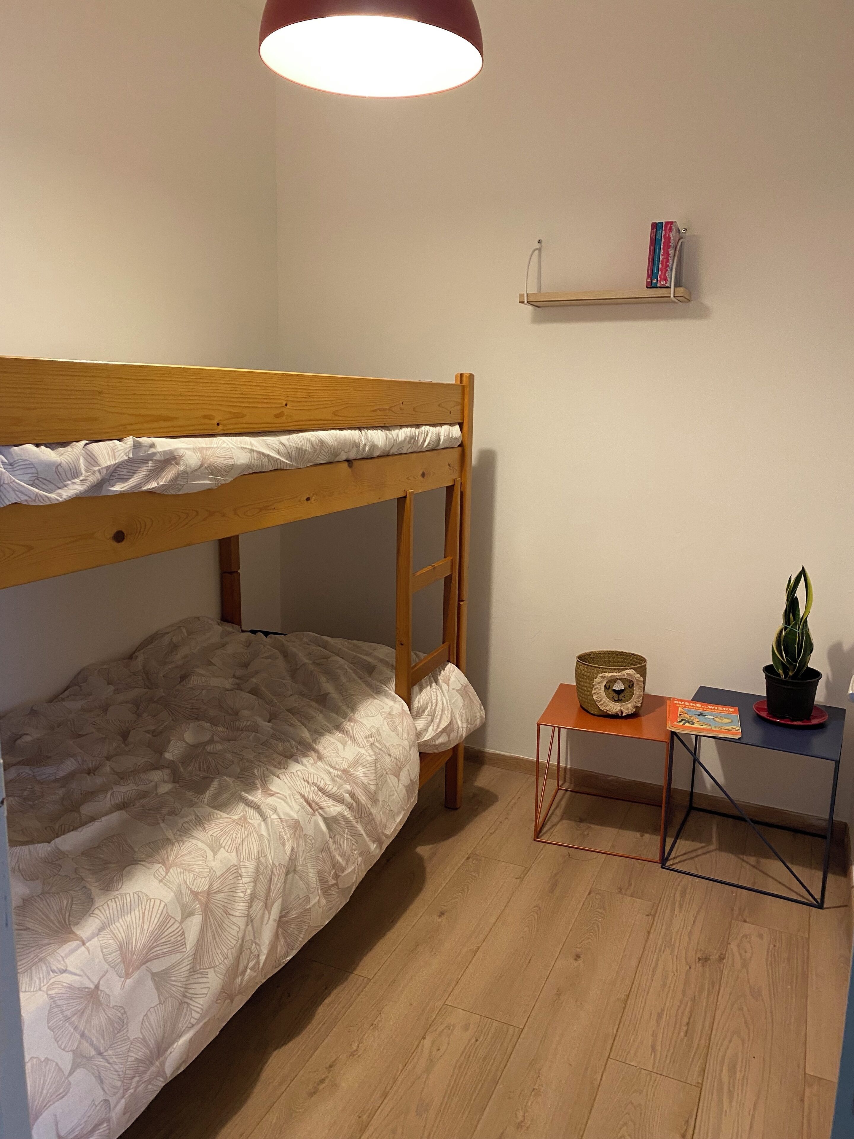 Una cama individual con estructura de madera, una colcha blanca con diseño, una mesita con una planta y un libro, y una estantería con libros y una cesta.
