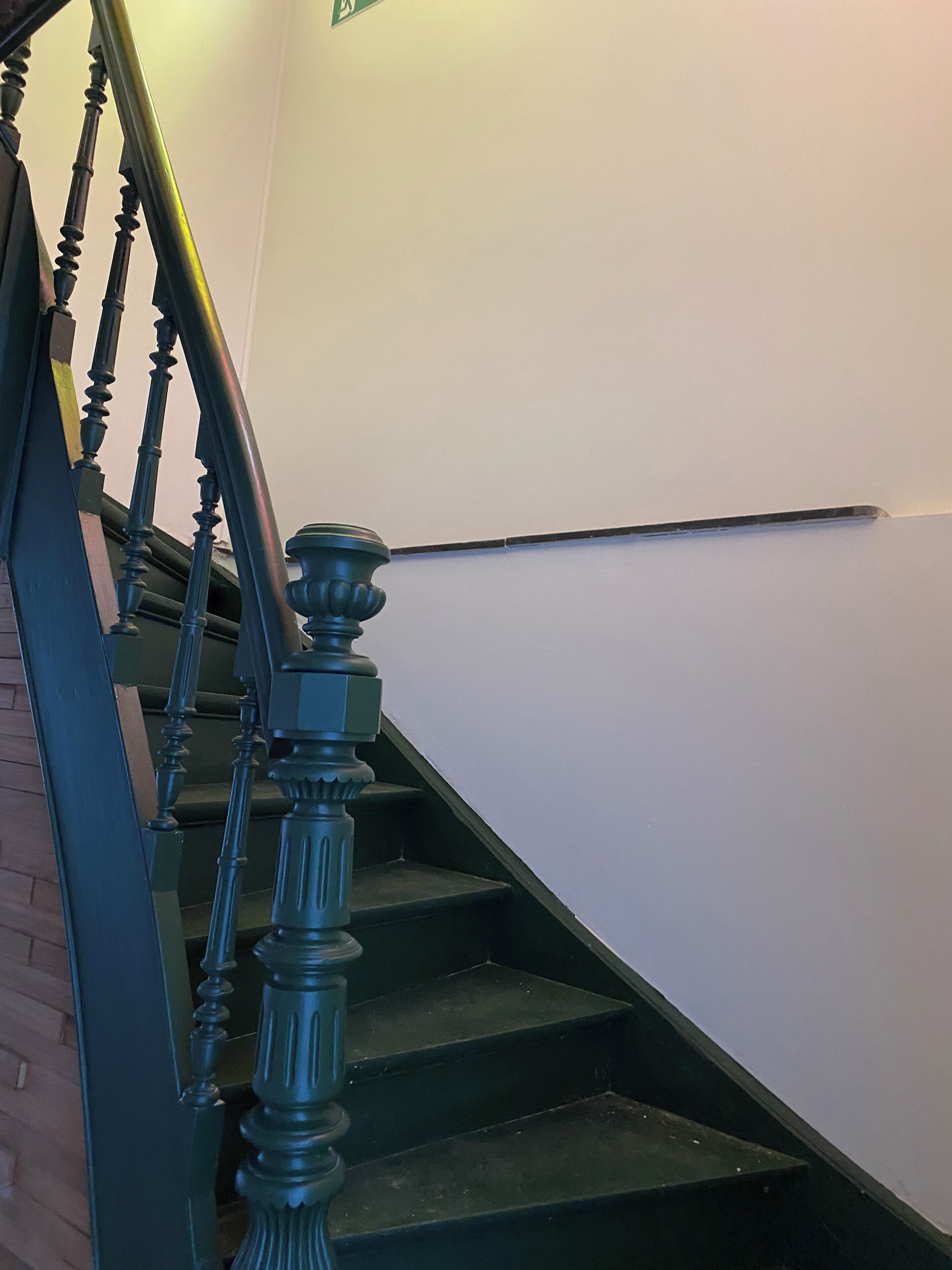 Una escalera con balaustres de madera oscura y barandilla, que lleva a un rellano con pared blanca y un letrero de salida de emergencia verde.