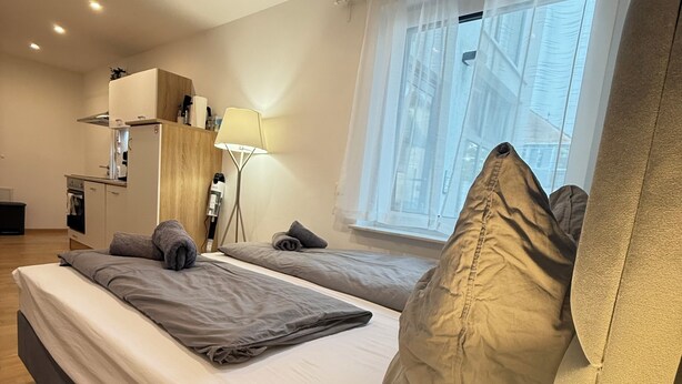 Une chambre d’hôtel moderne avec un grand lit, une table de chevet, une lampe, une petite kitchenette et une fenêtre donnant sur des immeubles.