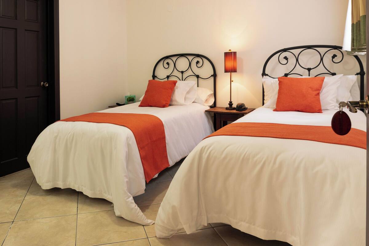 Two single beds with white and orange bedding, a nightstand with a lamp, and a door in the background.