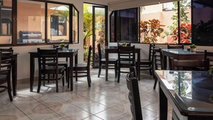 A dining area with multiple tables and chairs, large windows with views of plants and a building outside. - Brisas del Aeropuerto (La Asunción)