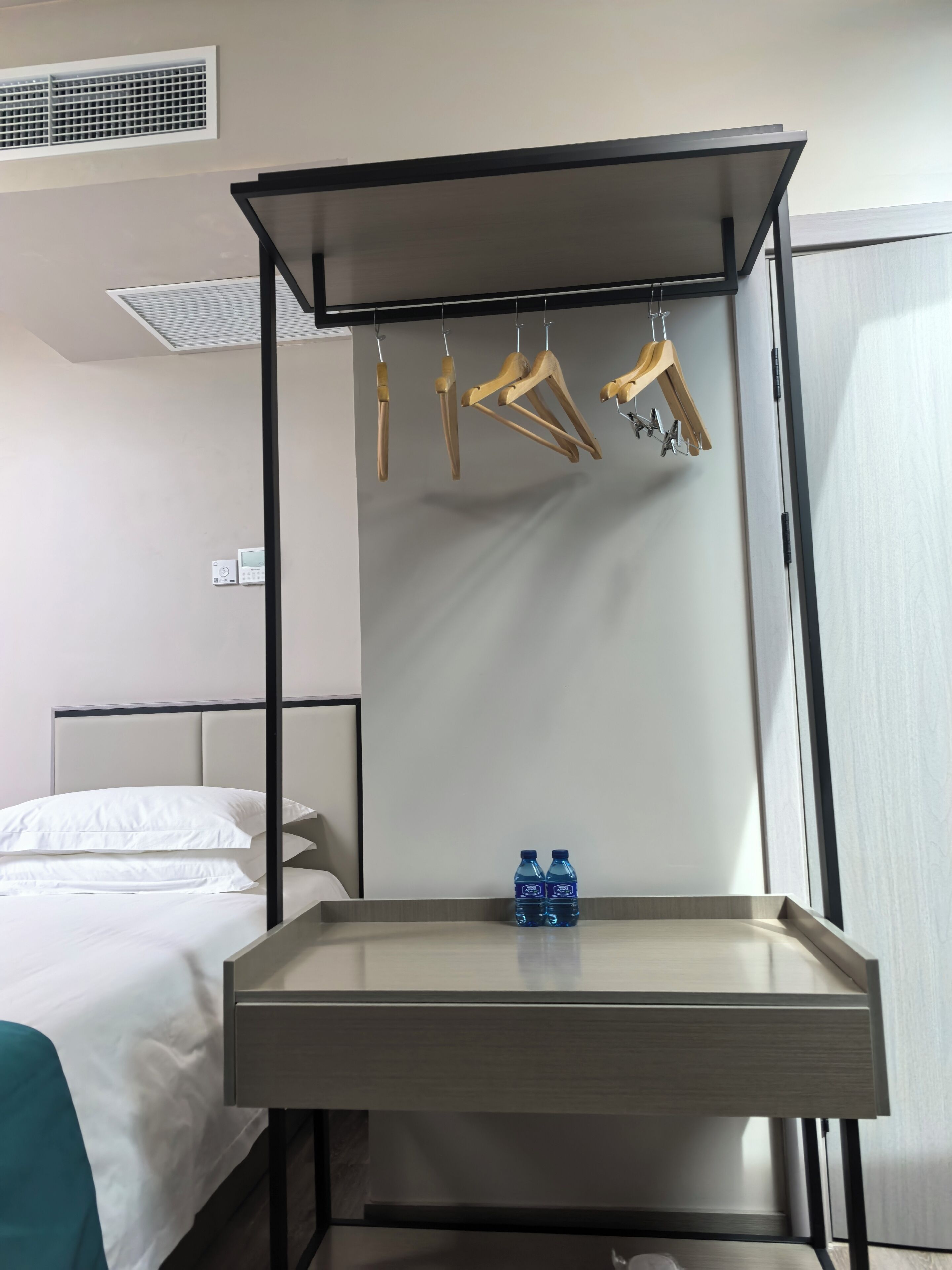 A hotel room with a bed, a bedside table with two water bottles, a hanging clothes rack, and a thermostat on the wall.