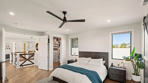 A modern bedroom with a large bed, a ceiling fan, a nightstand, a window with a view, and a walk-in closet.