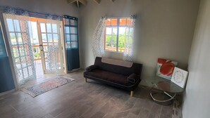 A room with a dark brown sofa, a glass coffee table, and a rug. There is a window with patterned curtains and a view of greenery outside. - Home in Carellie Gardens (Bisee)