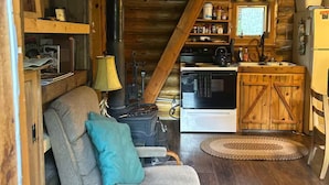 A cozy cabin interior with a wooden ceiling, a sofa with cushions, a fireplace, a kitchen area with a stove and refrigerator, and a window with a view. - Walden Cabin Yellowstone (Silver Gate)