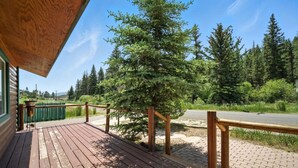 A wooden deck with a railing, a large evergreen tree, and a paved path leading to a road. - Stunning Log Cabin Close to Panguitch Lake (Panguitch)
