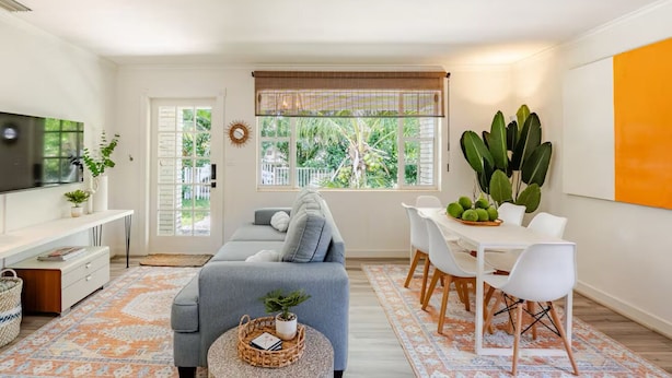 A modern living room with a grey sofa, dining table, and chairs, a TV, and large windows with blinds.