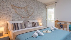 Une chambre avec un mur en pierre, un lit recouvert d’une couette bleue, des oreillers, une lampe de chevet, un berceau et une fenêtre avec des rideaux.