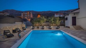 Une piscine entourée de chaises longues et bordée d’un mur de pierre, le tout niché au milieu de bâtiments et avec une chaîne de montagnes en toile de fond.