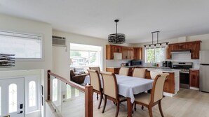 A modern kitchen with wooden cabinets, a dining area with a table and chairs, and a view of the outside through large windows. - Okanagan Escape (Kelowna)