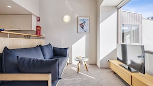 A modern living room with a dark blue sofa, a small round table with a magazine, a flat-screen TV on a wooden stand, and a large window offering a view of the outside.
