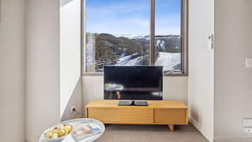 A modern room with a large window, a flat-screen TV on a wooden console, a small table with a bowl of fruit, and a magazine.