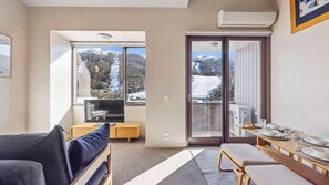 A modern living room with a sofa, a TV, a dining table with chairs, and large windows offering a view of snow-covered mountains.