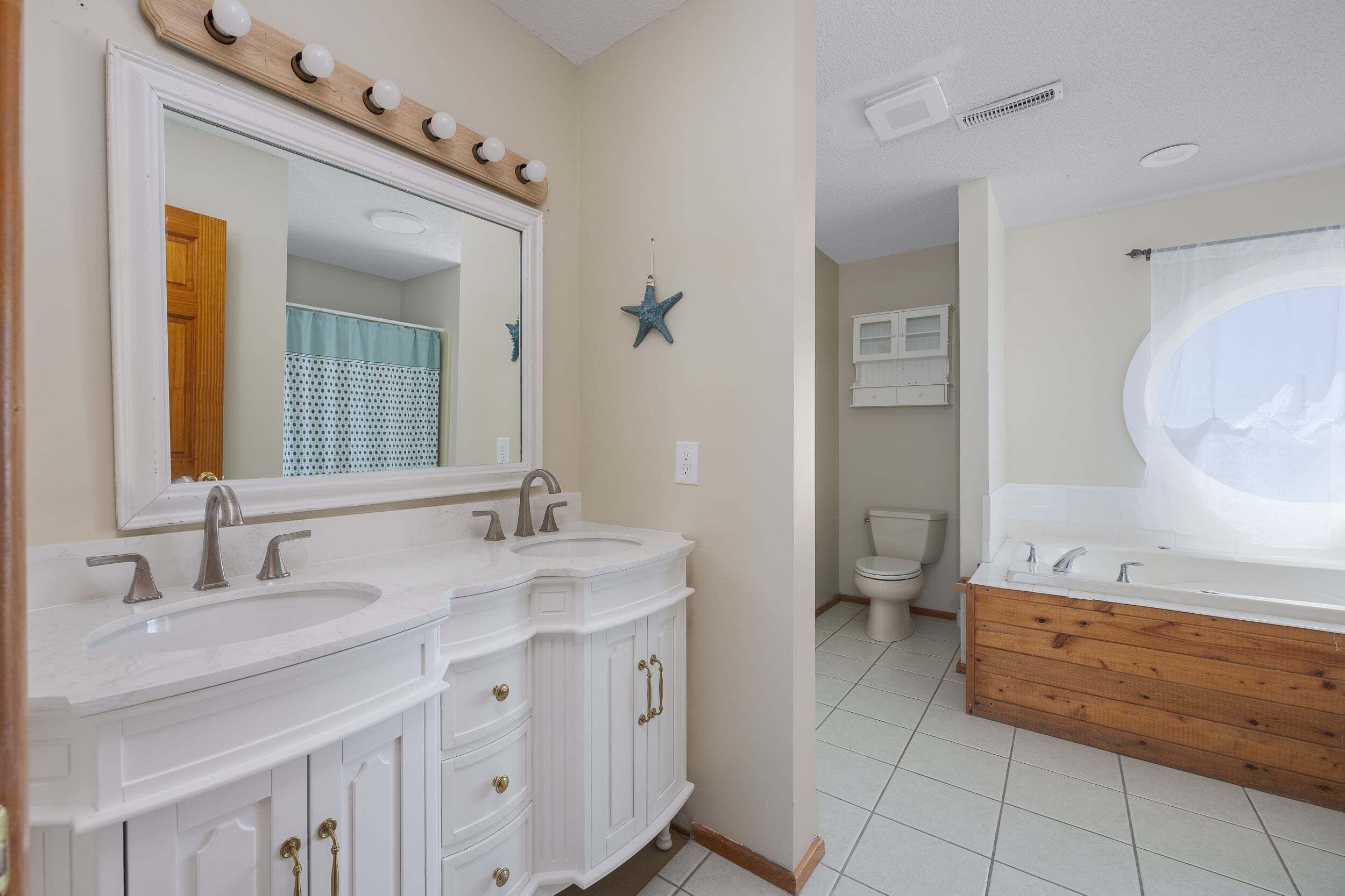 A bathroom with a double sink vanity, a large mirror with lights, a star decoration, and a bathtub.