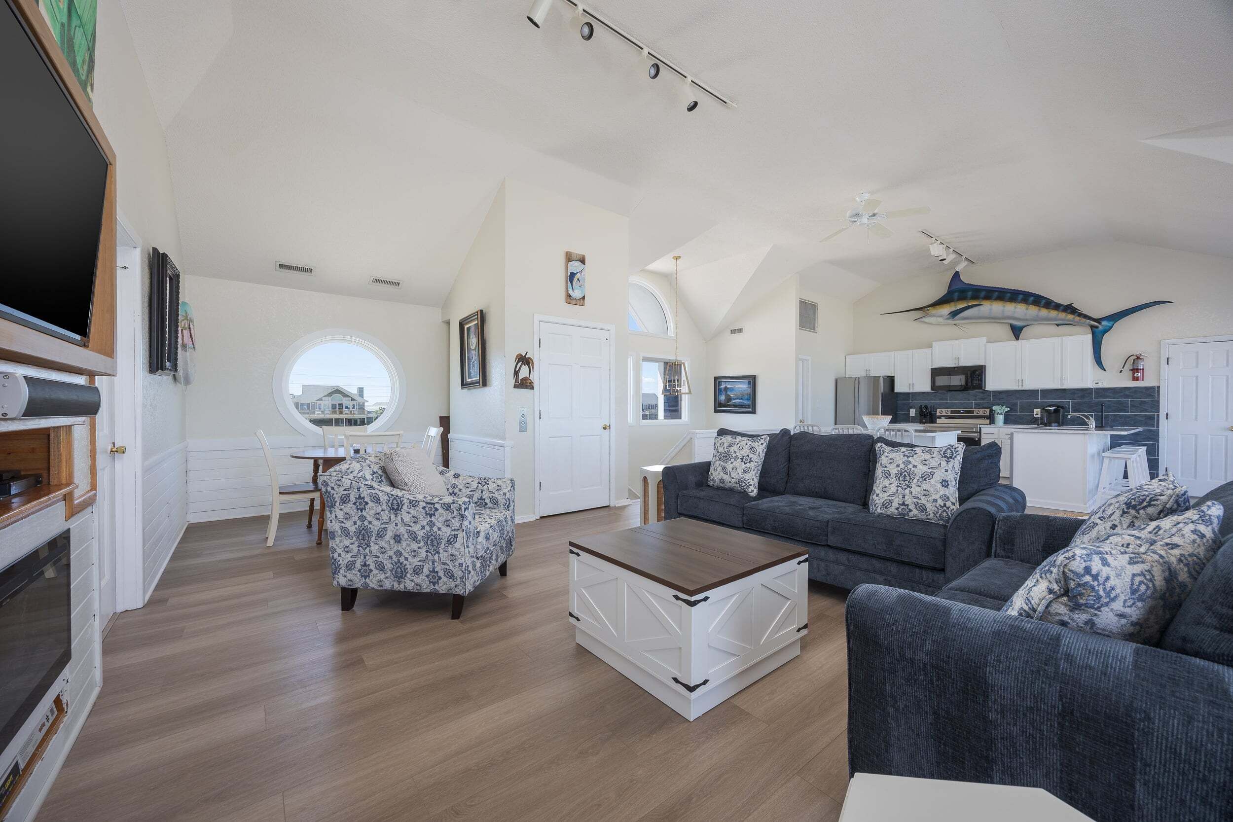 A spacious living area with a fireplace, a round window, a dining table, and a kitchen in the background.