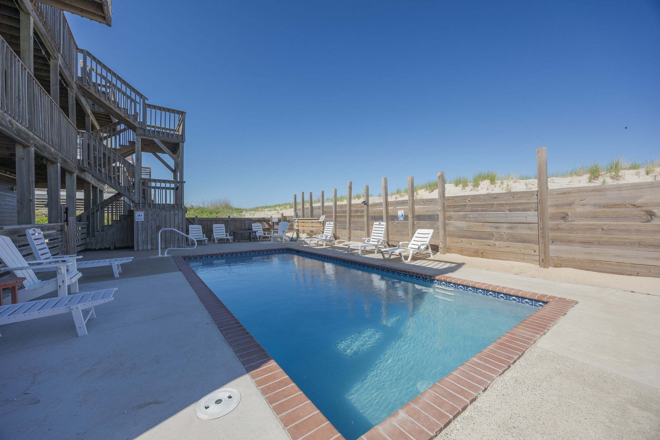 A swimming pool with lounge chairs, a wooden deck, and a sandy beach in the background.
