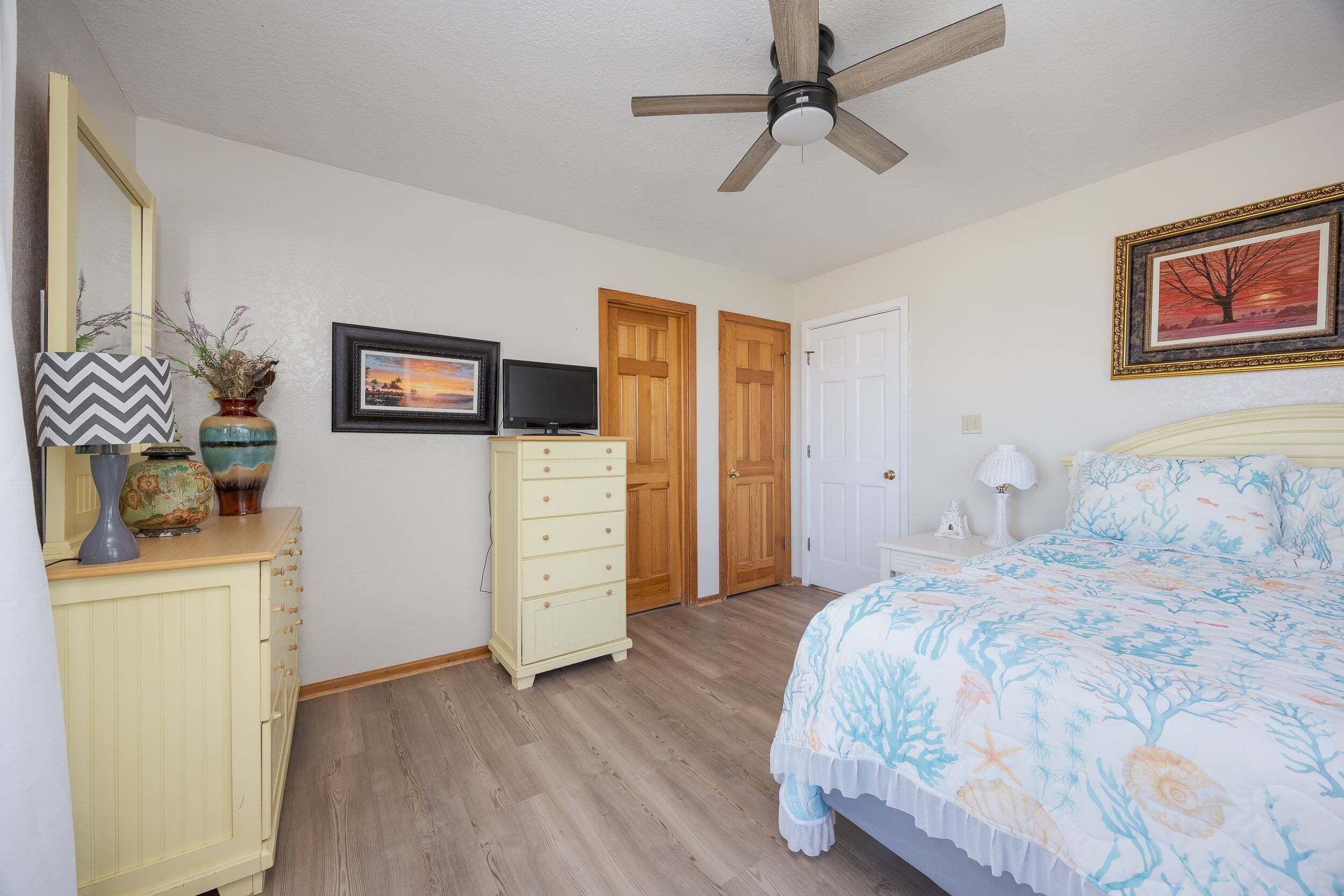 A bedroom with a bed, dresser, vanity, television, and two doors.