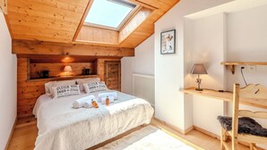 A cozy bedroom with a sloped ceiling, a skylight, a bed with white bedding, a bedside table with a lamp, and a shelf with a picture.