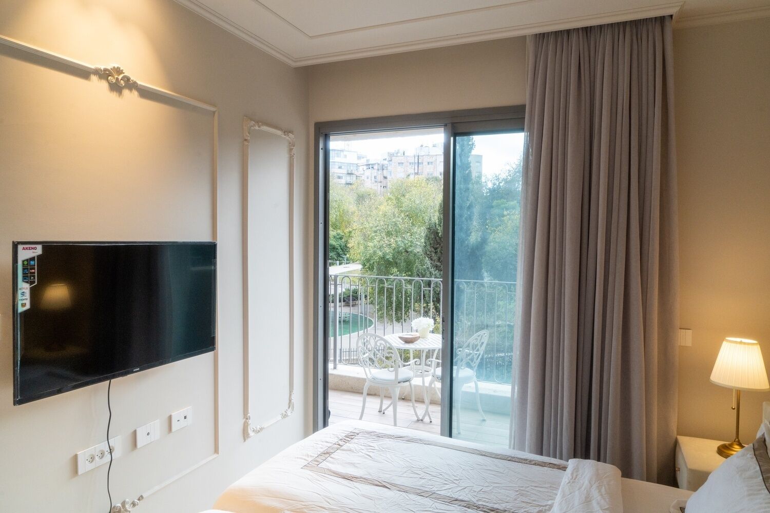 A hotel room with a bed, a TV mounted on the wall, a balcony with a table and chairs, and a view of greenery and buildings outside.