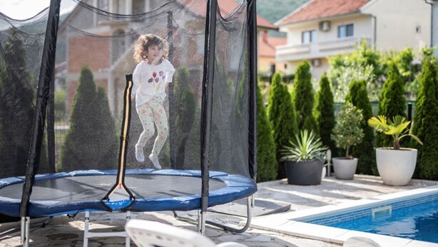 A trampoline with a safety net, a poolside lounge chair, and a pool with a clear blue water.