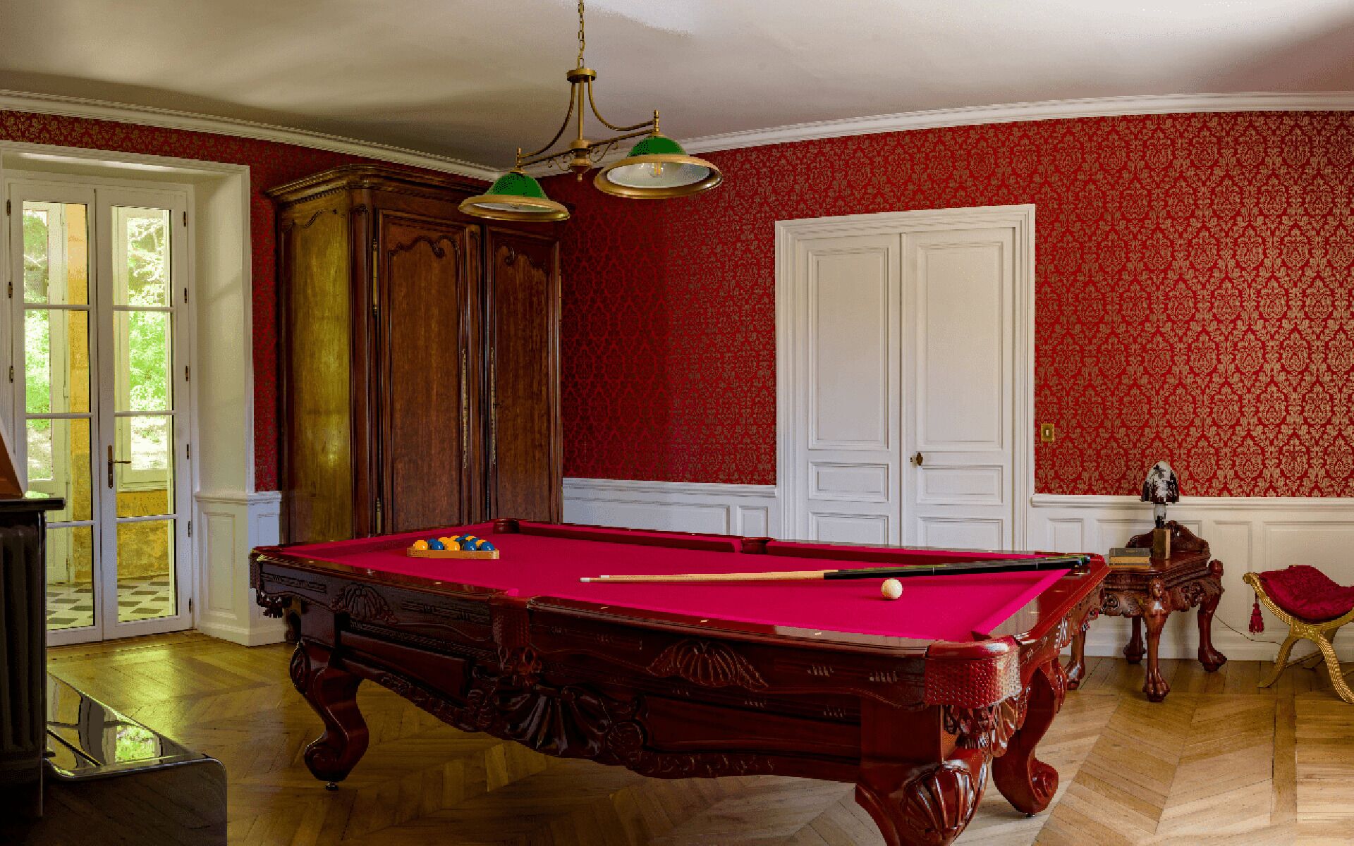 Een biljarttafel in een kamer met rood behang, houten kasten en een deur.
