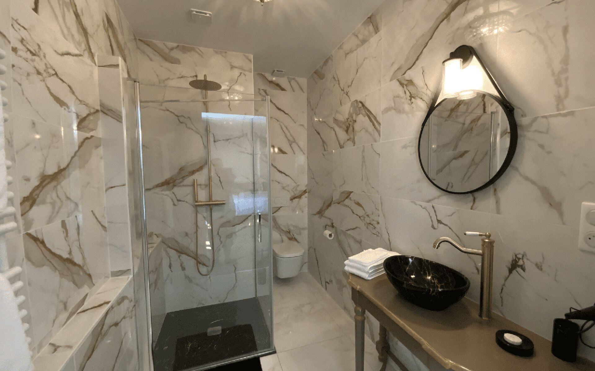 Une salle de bain dotée d’une douche à parois de verre, d’un mur en marbre, d’un miroir rond et d’un lavabo avec un receveur noir.