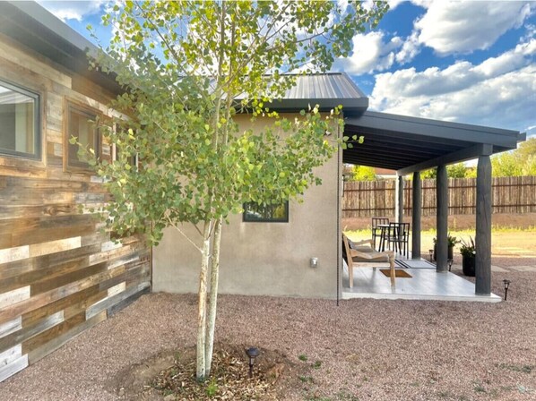 A modern house with a wooden exterior, a covered patio with outdoor furniture, and a tree in the yard. - NM Farmhouse Sanctuary w/soaking rm 1 acre private (Santa Fe)