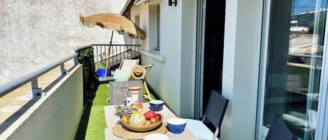 A balcony with a table set for dining, a chair, a thatched umbrella, and a window.