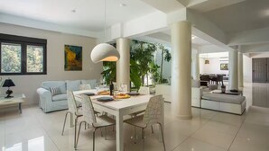 A spacious living area with a dining table, white sofas, and a large window with a view of greenery.