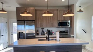 Una cocina moderna con una isla central, electrodomésticos de acero inoxidable, armarios de madera, iluminación colgante y encimera negra.