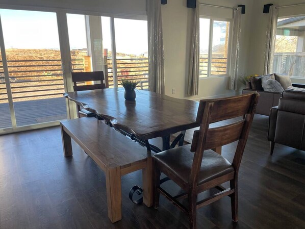 Un comedor con mesa y sillas de madera, ventanas amplias con vista a un paisaje árido y un sofá al fondo.