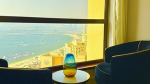 A room with a view of a beach and cityscape, a round table with a vase, and two blue chairs.