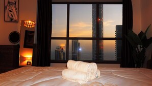 A hotel room with a large window, a bed with neatly folded towels, a lamp, and a framed picture on the wall.