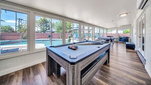 A pool table in a room with large windows, a pool, and a patio area.