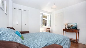 Une chambre à coucher avec un lit, une table de chevet en bois avec une lampe, une télévision et une fenêtre donnant sur un jardin.
