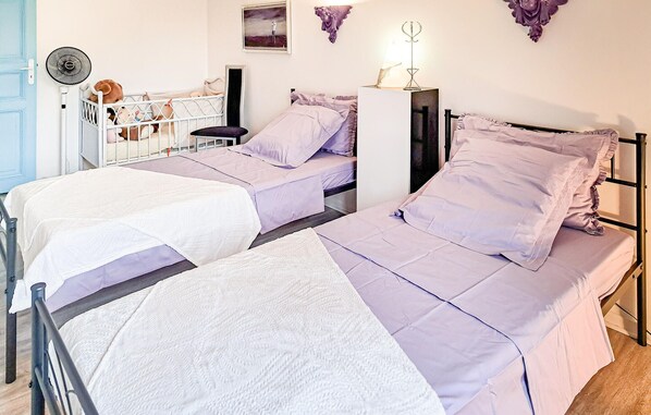 Une chambre à coucher avec deux lits, chacun recouvert d’une couvre-lit blanche et violette. On y trouve un radiateur blanc, une petite table avec une lampe et une étagère murale ornée d’objets décoratifs.