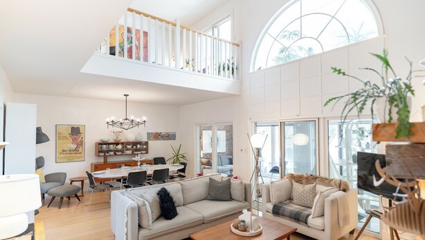 A spacious living room with a high ceiling, a dining area, and a staircase leading to an upper level.