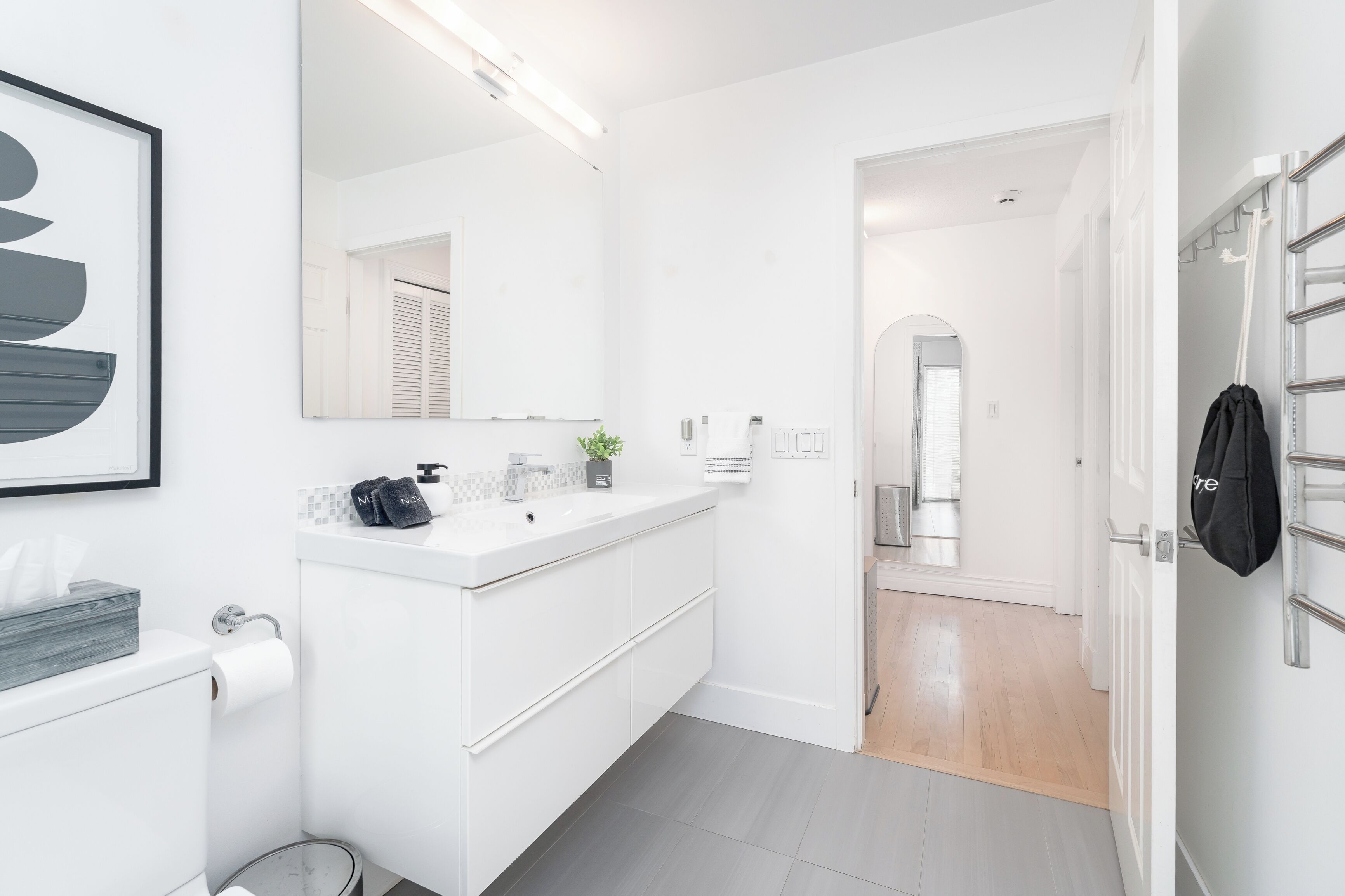 A modern bathroom with a white sink, mirror, and toilet. There is a towel rack and a small plant on the counter.