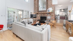 A spacious living room with a fireplace, a kitchen in the background, and a large window providing natural light.