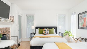 A modern bedroom with two beds, a fireplace, a round table, and a large window.