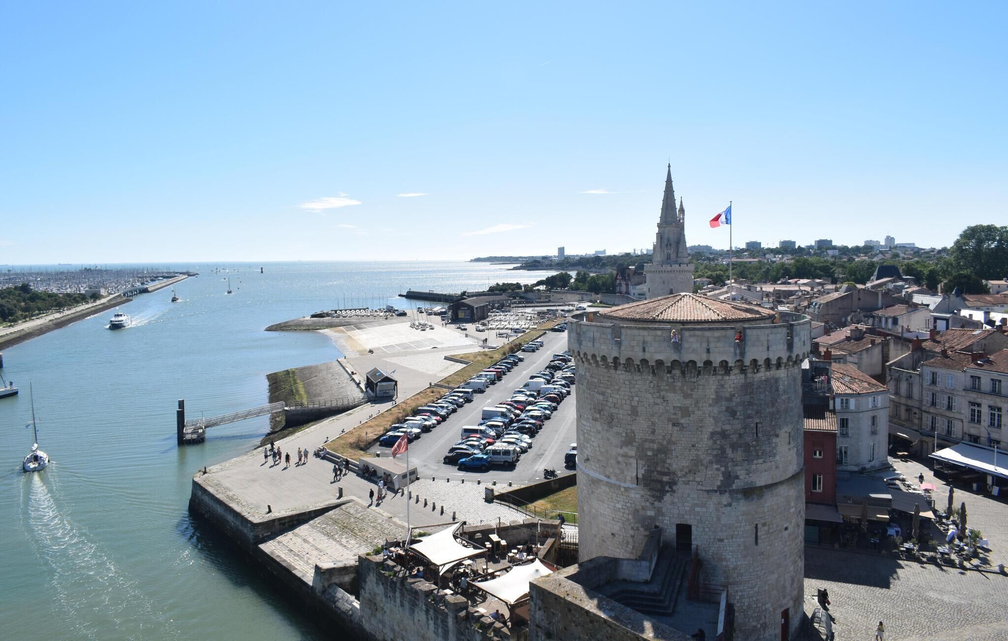 Paisagem costeira com uma torre imponente, um porto com barcos e uma área de estacionamento.