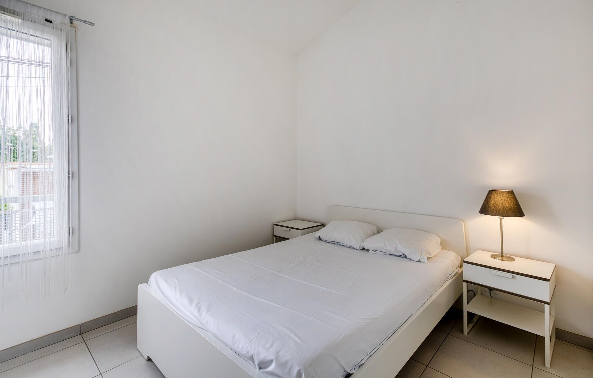 Une chambre à coucher au style minimaliste, avec un lit simple, une table de chevet, une lampe et une fenêtre avec des rideaux.