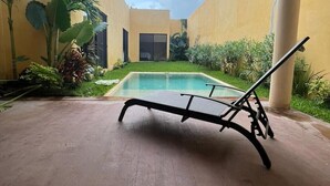 Zona de piscina con una tumbona, rodeada de vegetación y un edificio de fachada amarilla.