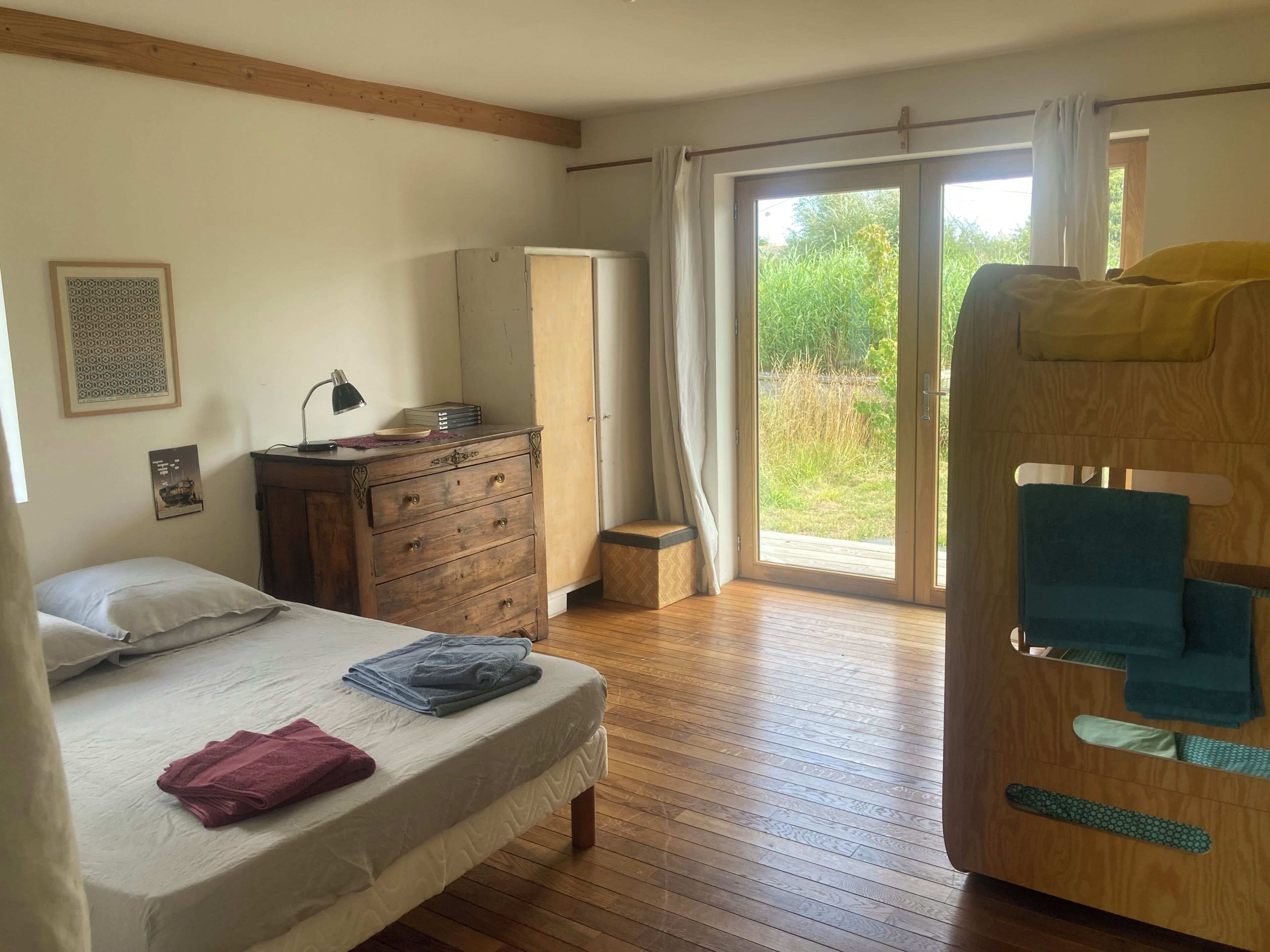 Un dormitorio con cama, cómoda de madera, escritorio con lámpara y puerta corrediza de vidrio que da a un espacio exterior.