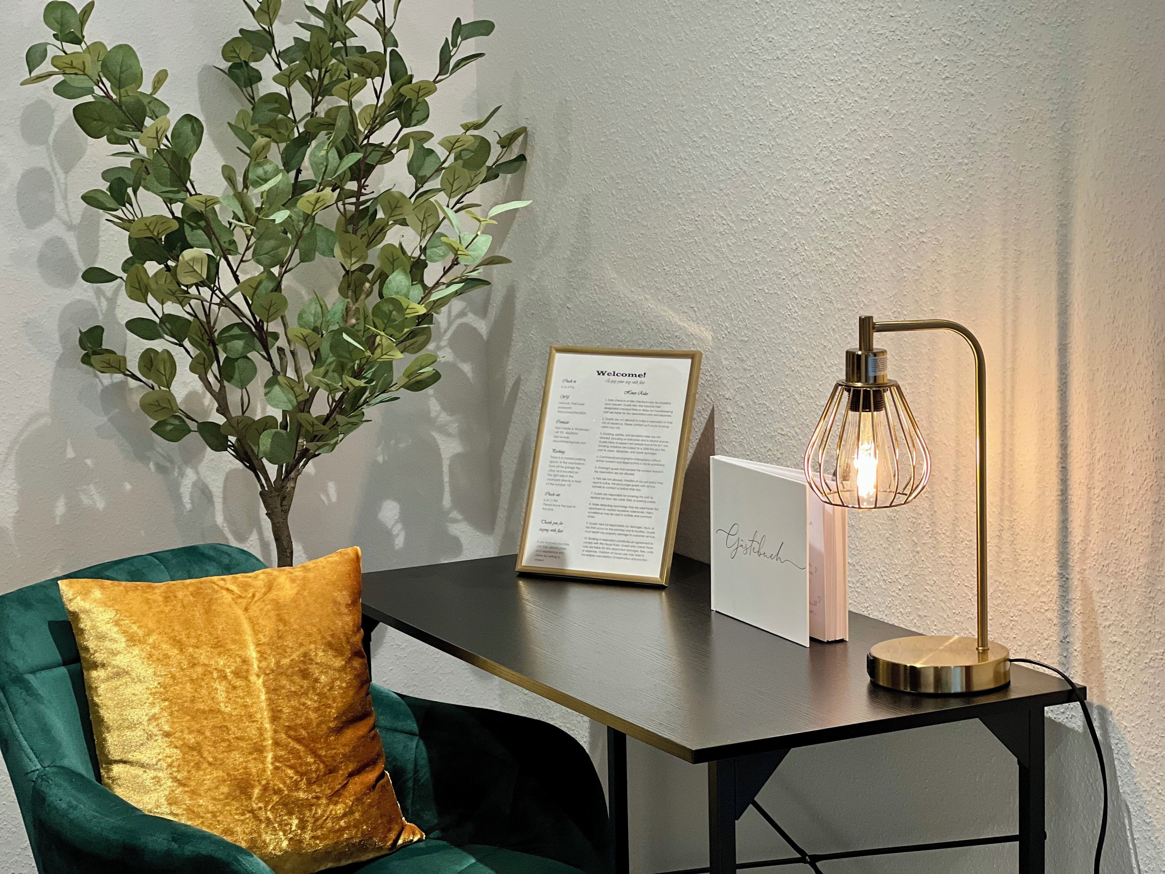 Een kamer met een groene fluwelen bank, een gouden sierkussen, een klein tafeltje met een bord 'welkom', een decoratieve lamp en een pot met een plant.