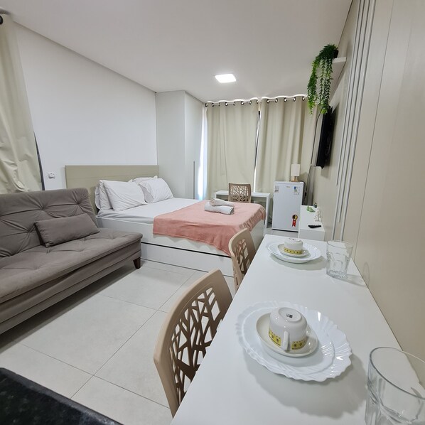 Una habitación de hotel moderna con cama, sofá, mesa de comedor con tetera y tazas, y un refrigerador.