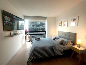 A bedroom with a bed, bedside tables, a TV, and a window with a city view. - Loft con Tecn- Alexa (Bogotá)